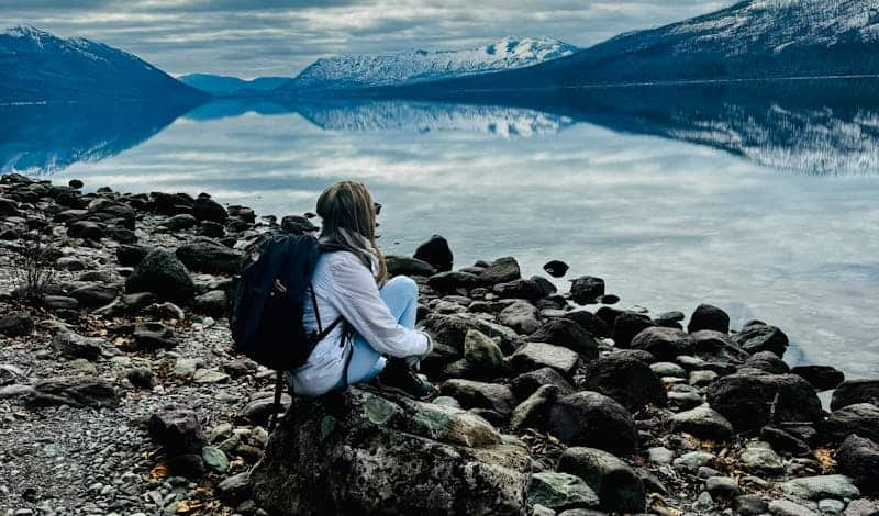 A woman in warm clothing sits by a tranquil mountain lake, reflecting the surrounding landscape.