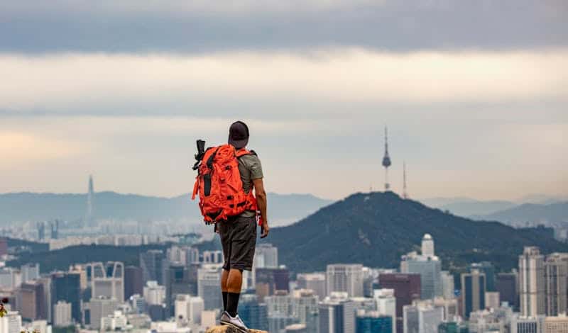 Man with backpack observing Seoul cityscape from mountain peak.